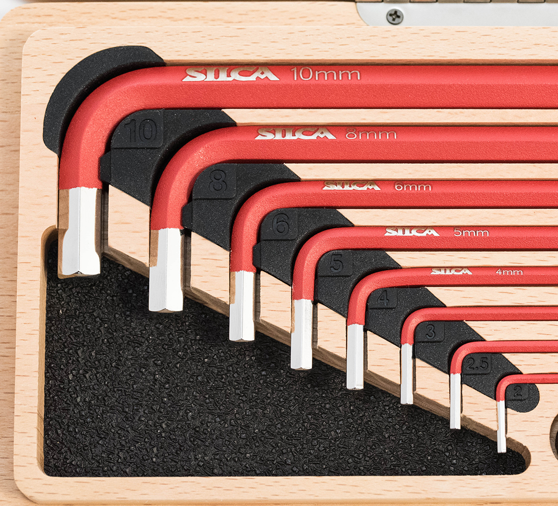 Object: red L-shaped hex (Allen) keys. Action: neatly nested by size with tips pointing downward and millimeter labels visible. Context: fitted into a wooden tray with a black foam insert.