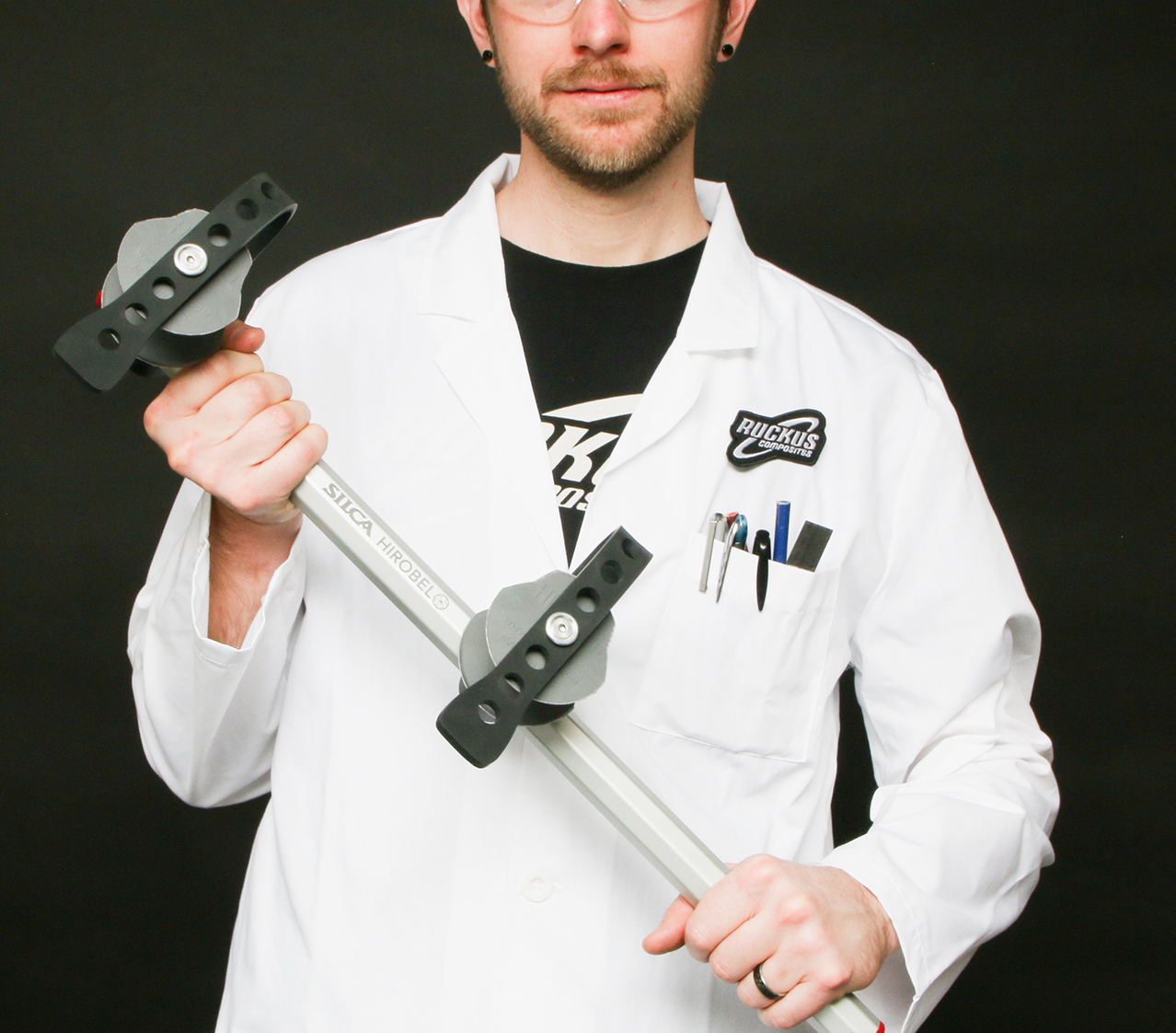 Person in a white lab coat holds a long metal bar with two black adjustable clamps horizontally across their chest against a dark background.